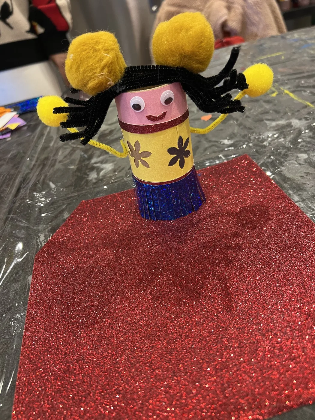 Tube doll with pom-pom arms on red glitter