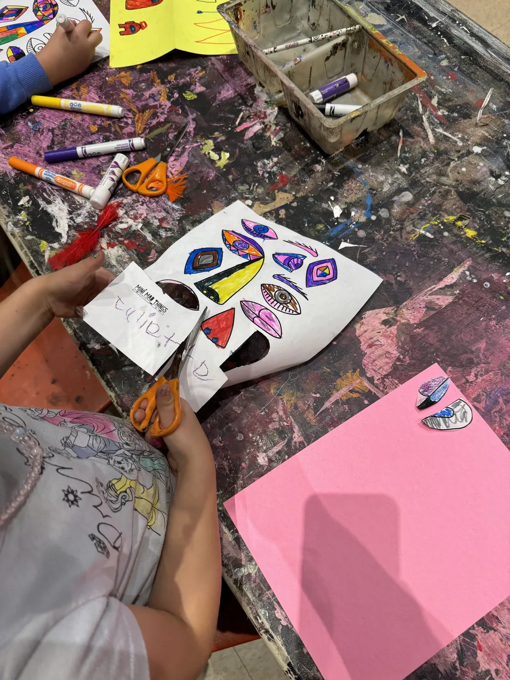 Hands cutting out coloured eye shapes on pink paper