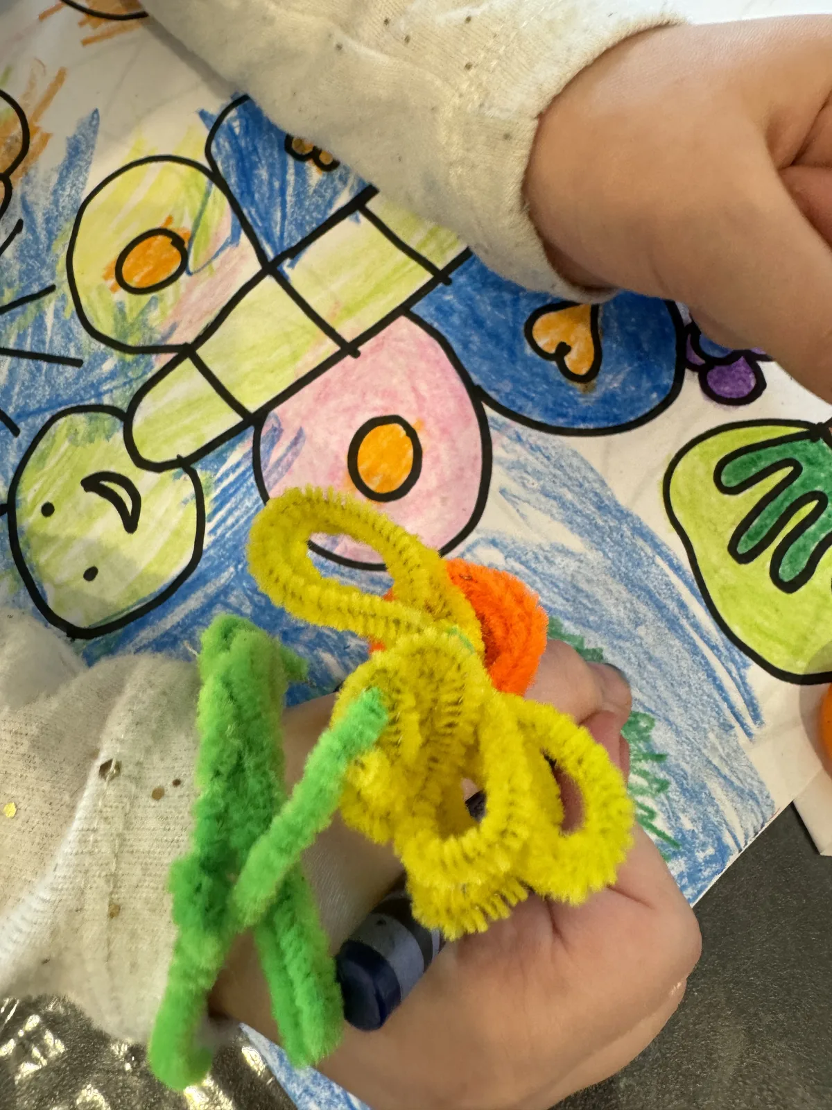 A child's hands holding blue and green pipe cleaners over a drawing