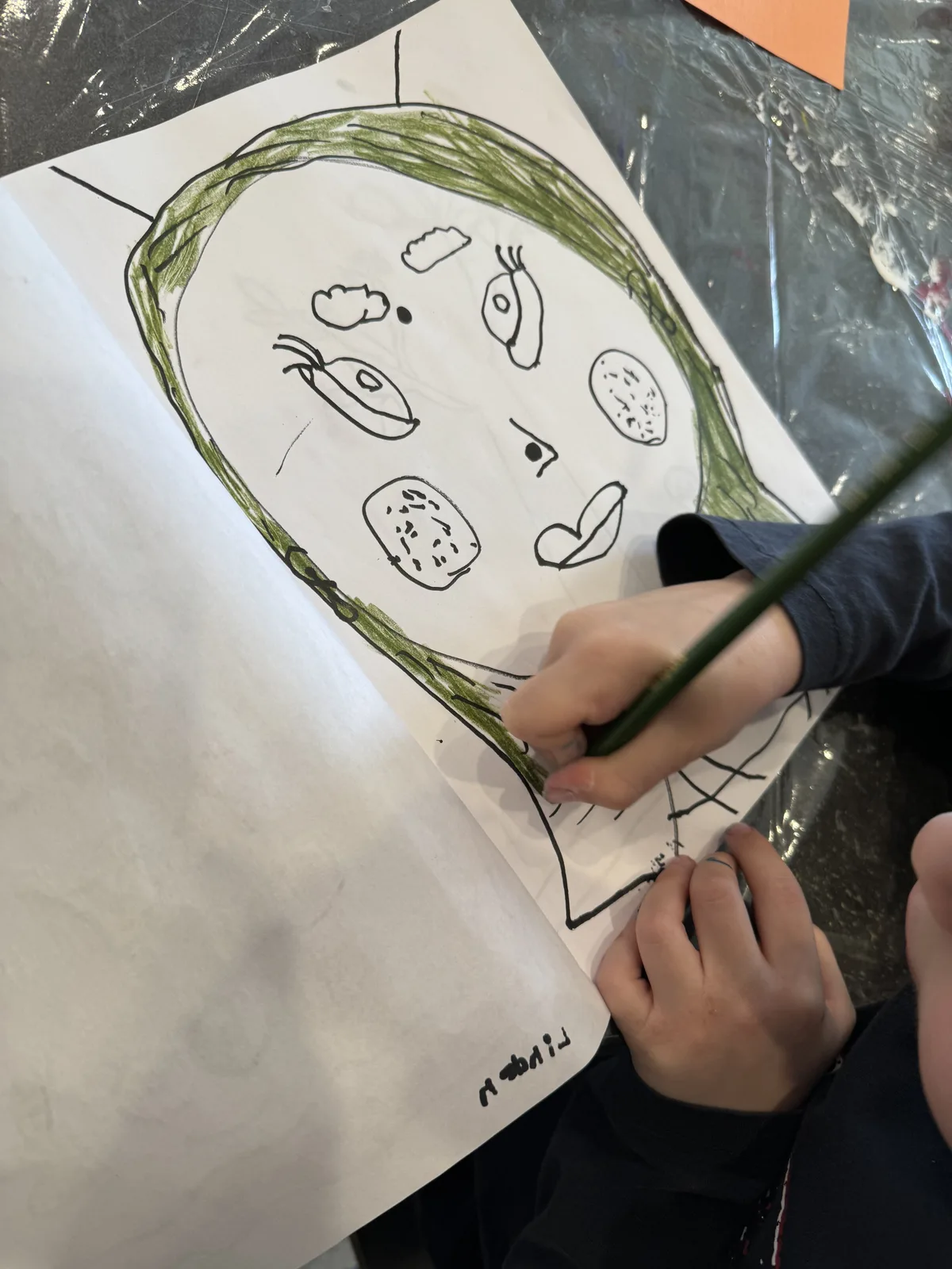 A child's hands drawing green hair onto a face-outline sketch