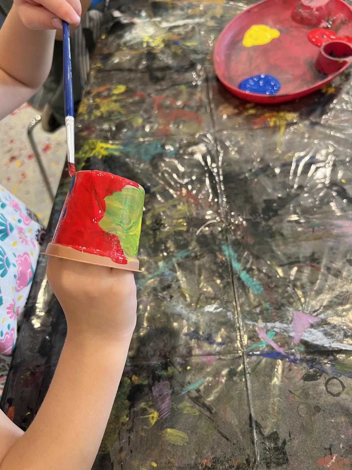 A child's hand painting a clay pot in red and green