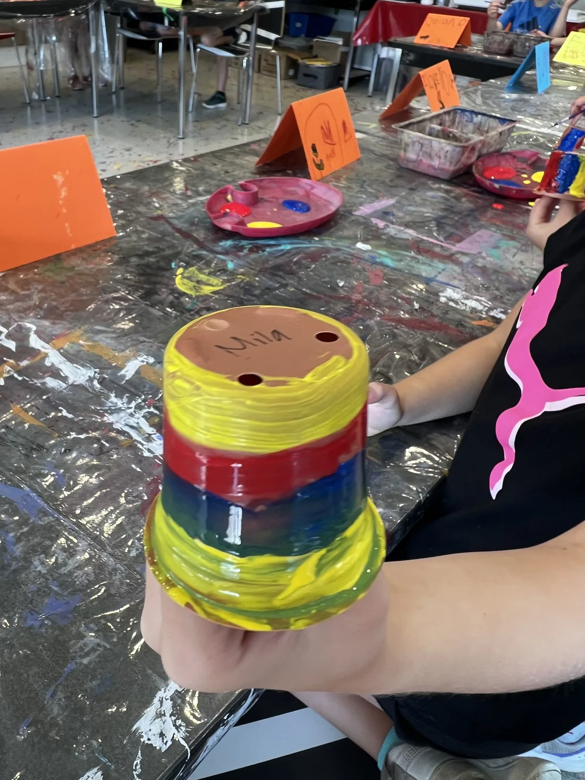 A child holding a rainbow-painted pot labelled "Mila"
