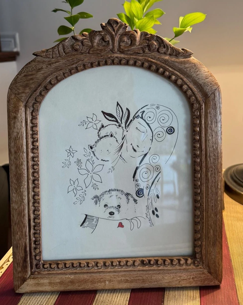 A carved wooden frame on a client’s table holding an ink drawing of a woman with two small dogs and a red heart