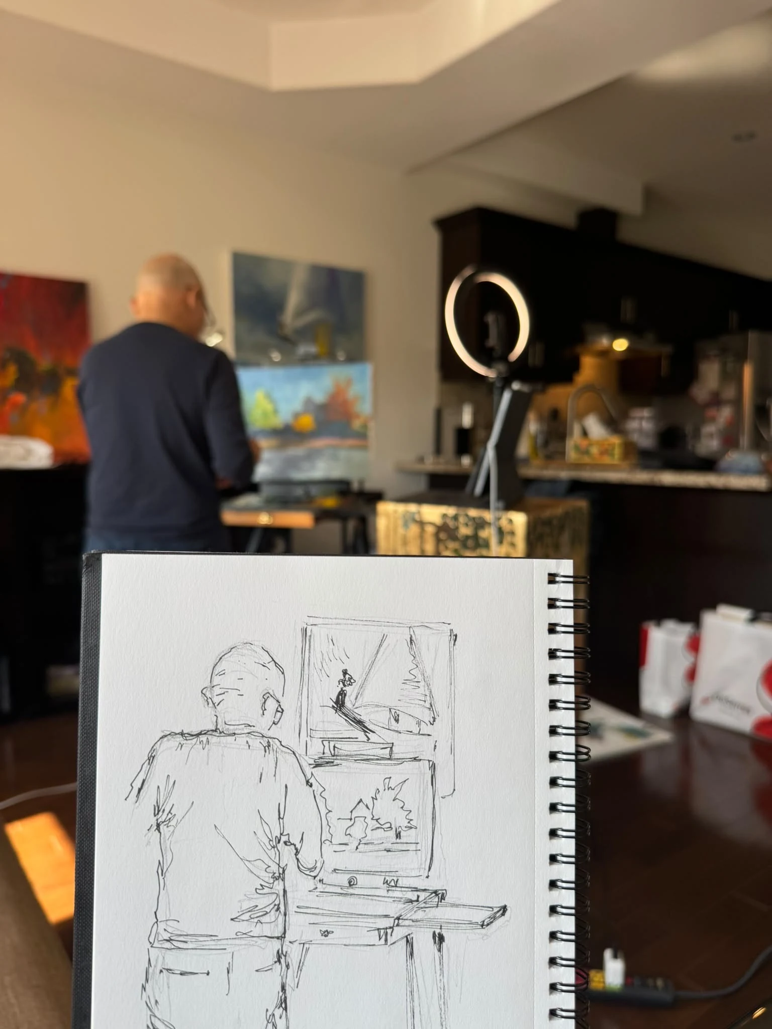 Aroub’s open sketchbook held up in front of a friend painting at his easel: the ink sketch of his back and the easel, with the real scene behind